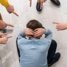 Chłopiec skulony na podłodze, otoczony przez palce wskazujące na niego. To obraz tego, czym jest bullying.