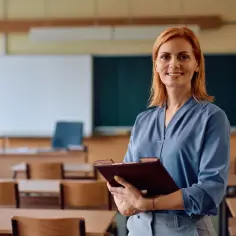 Uśmiechnięta nauczycielka z rudymi włosami w klasie, trzymająca teczkę. To inspiracja dla tych, którzy zastanawiają się, jak zostać nauczycielem.