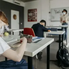 Uczniowie w maseczkach siedzą w klasie. Jedna dziewczyna czyta książkę, inny uczeń śpi. Nauczycielka stoi przy tablicy. EDB co to?
