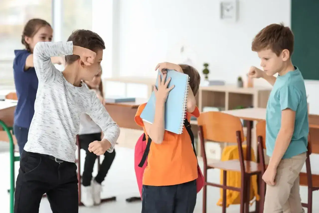 Dzieci w klasie. Jeden chłopiec grozi drugiemu, który zasłania się zeszytem. To jest bullying, co to znaczy?