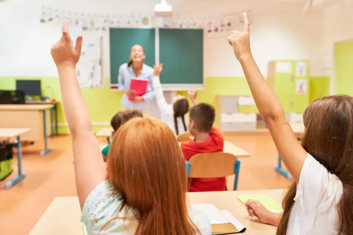 Uczniowie z podniesionymi rękami, ciekawi, czy edukacja zdrowotna będzie obowiązkowa. Nauczycielka z uśmiechem czeka na odpowiedzi.