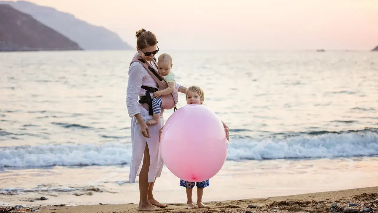Mama z dziećmi na plaży, maluch w nosidle, starszak z balonem. Radosne chwile i bezpieczne wakacje nad morzem.