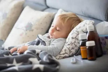 Małe dziecko śpi otulone kocykiem. Obok leżą buteleczki z lekami, w tym antybiotyk dla dzieci, oraz chusteczki.