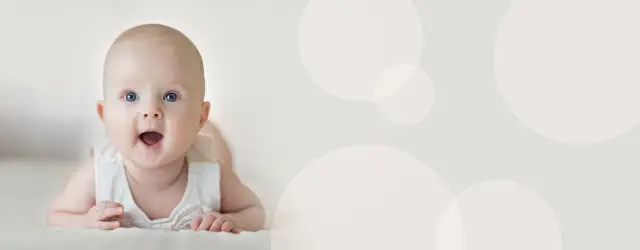 Uśmiechnięte, 4 miesięczne dziecko z szeroko otwartymi oczami i buzią leży na brzuchu.
